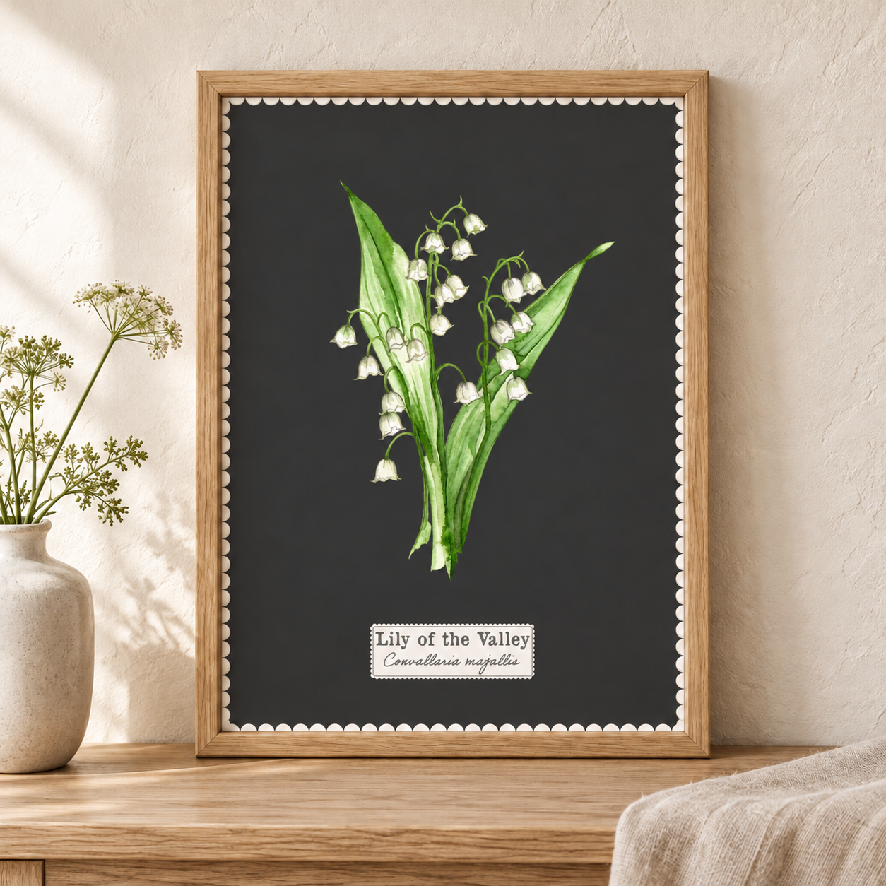 A framed botanical art print of a lily of the valley on a dark charcoal background, leaning on a wooden table against a textured beige wall with a vase of flowers nearby.
